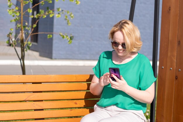 Portrait d'une étudiante souriante à lunettes de soleil tenant un smartphone dans ses mains assise sur un banc dans un parc de la ville