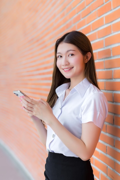 Portrait d'une étudiante thaïlandaise asiatique en uniforme sourit joyeusement en utilisant un smartphone