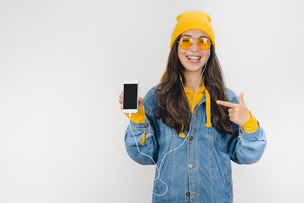 Portrait excité fille au chapeau et écouteurs pointant le doigt sur un téléphone mobile à écran blanc