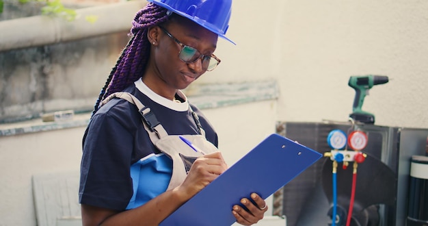 Portrait d'un expert afro-américain souriant faisant l'inspection des niveaux de réfrigérant et les réparations nécessaires pour éviter les pannes majeures. Travailleur vérifiant le système hvac, écrivant les résultats sur le presse-papiers