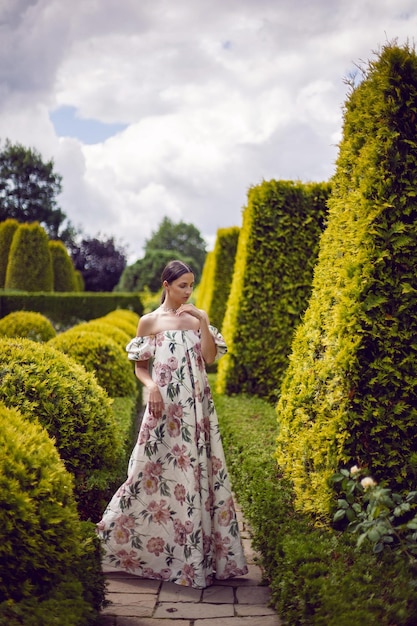 Portrait extérieur d'une belle femme brune de luxe vêtue d'une robe à fleurs se dresse dans un parc aux arbres taillés