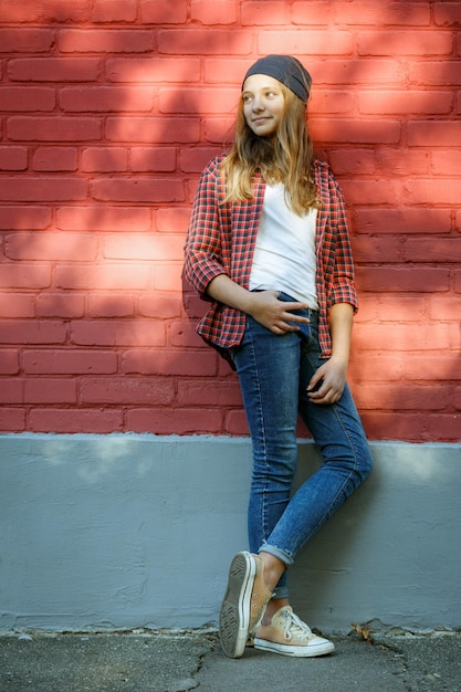 Portrait extérieur vertical de style de vie d'une jeune adolescente souriante portant des baskets, un chapeau en coton bleu et une chemise à carreaux rouge sur un fond de mur de briques rouges