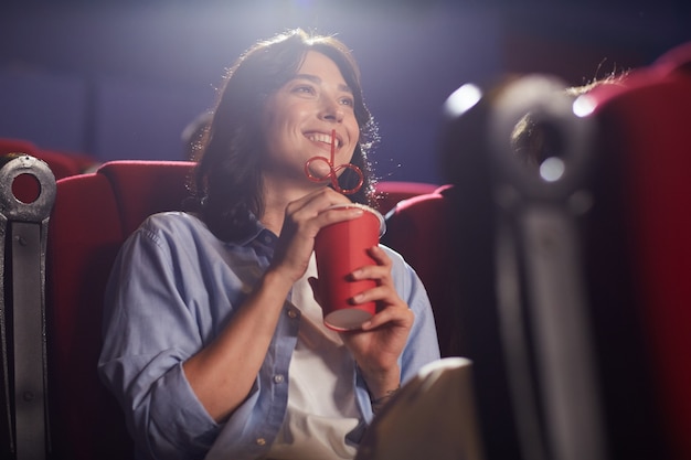 Portrait de faible angle de jeune femme souriante au cinéma regarder un film seul et boire du soda, regardant l'écran, copiez l'espace