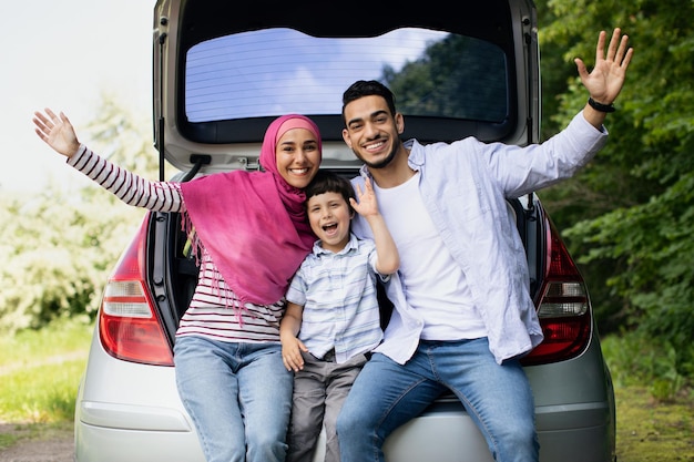 Portrait d'une famille arabe heureuse avec un petit fils assis dans un coffre de voiture et souriant à la caméra, des parents musulmans enthousiastes et leur enfant de sexe masculin appréciant de voyager ensemble avec une automobile, espace libre