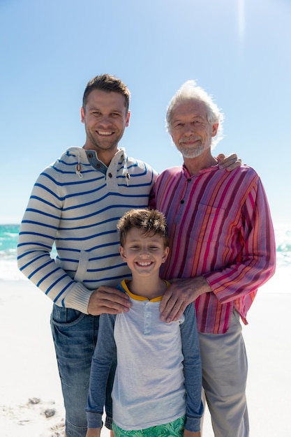 Portrait d'une famille caucasienne de plusieurs générations debout sur la plage avec le ciel bleu et la mer en arrière-plan, embrassant et souriant à la caméra