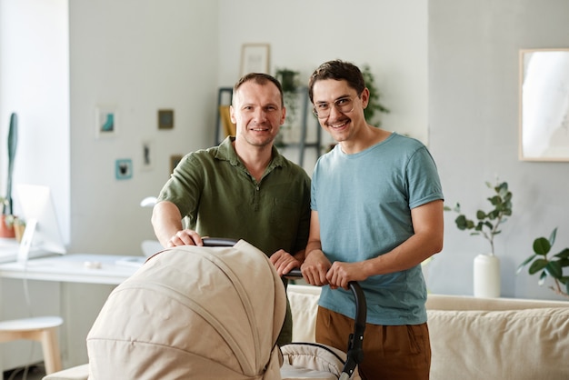 Portrait d'une famille gay heureuse debout dans la chambre avec une poussette de bébé et souriant à la caméra
