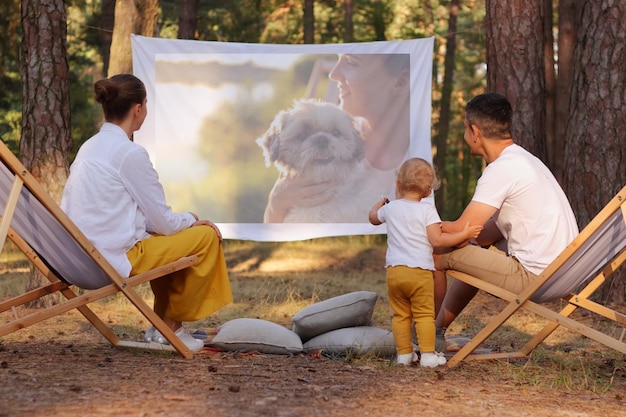 Portrait d'une famille heureuse assise dans la forêt sur des chaises longues avec leur petite fille et regardant un film sur des parents de projecteur jouant avec leur enfant mignon et profitant de la nature et de l'air frais