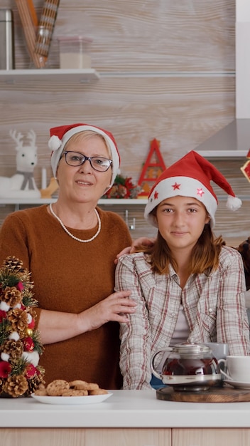 Portrait d'une famille heureuse portant un bonnet de noel regardant la caméra debout à table dans une cuisine décorée de Noël. Grands-parents profitant de la saison d'hiver passant des vacances de Noël avec leur petit-enfant