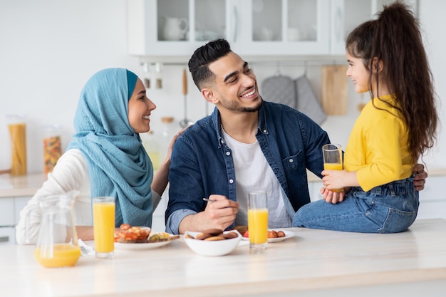 Portrait d'une famille musulmane heureuse de trois mangeant le déjeuner dans la cuisine ensemble, maman islamique, papa et enfant assis à table, appréciant une cuisine savoureuse à la maison et souriant, parents aimants regardant leur enfant