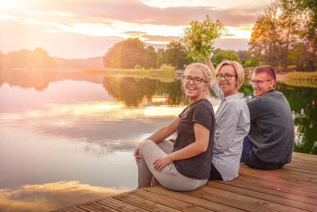 Photo portrait d'une famille souriante assise sur une jetée au-dessus du lac contre le ciel au coucher du soleil