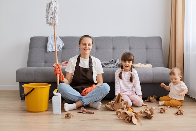 Portrait d'une famille souriante heureuse jeune adulte femme portant un tablier tenant une vadrouille dans les mains assis sur le sol près du canapé avec ses enfants regardant la caméra avec le sourire