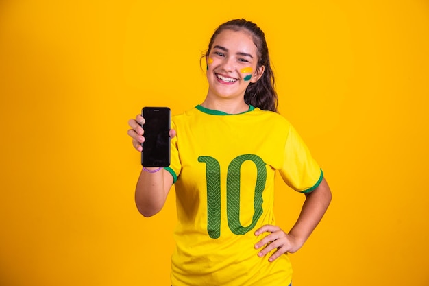 Portrait d'un fan brésilien Fan brésilien montrant son téléphone portable habillé en fan de football ou match de football sur fond jaune Couleurs du BrésilCoupe du monde