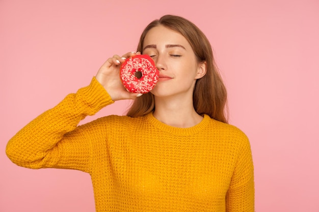 Portrait d'une fascinante fille au gingembre affamée en chandail sentant un beignet et exprimant le désir de manger une tentation de beignet sucré de confiserie sucrée prise de vue en studio isolée sur fond rose