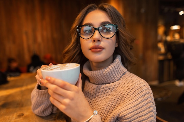 Portrait féminin à l'intérieur d'une belle fille à la mode avec des lunettes à la mode dans un élégant pull beige vintage tricoté tient une tasse blanche et boit du café dans un café