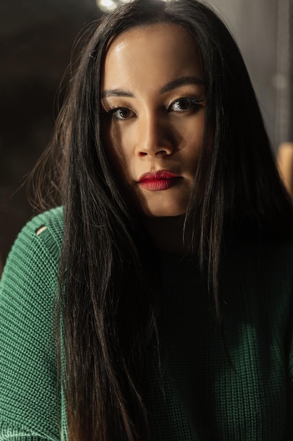 Portrait féminin d'une jolie jeune femme vietnamienne élégante avec du maquillage dans un pull en tricot vintage à la mode regarde la caméra dans une pièce dans l'obscurité