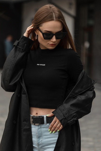 Portrait féminin à la mode d'un beau modèle de femme à la mode avec des lunettes de soleil cool en long manteau noir en cuir avec un t-shirt noir et un jean se promène dans la rue Mode et beauté des vêtements pour femmes urbaines