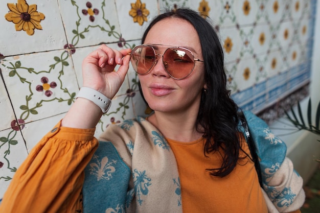 Portrait féminin de mode d'une belle fille élégante dans une robe jaune vif avec une écharpe verte vintage portant des lunettes de soleil rondes sur un fond de tuiles portugaises rétro