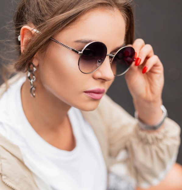 Portrait féminin à la mode d'une belle jeune fille avec des lunettes de soleil vintage et des lèvres roses dans un t-shirt blanc à la mode et un coupe-vent dans la rue Sports mode et beauté de style rétro