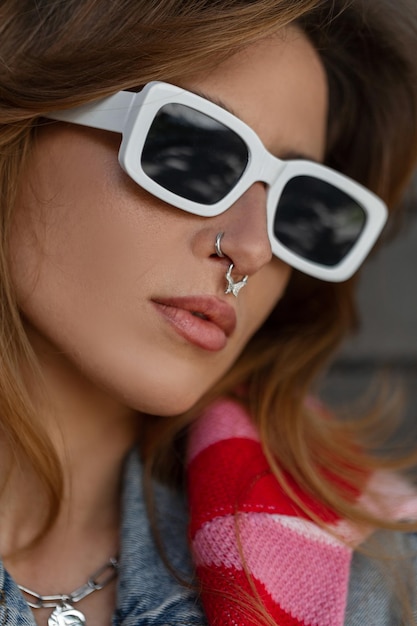 Portrait féminin de mode en gros plan d'une jolie jeune fille hipster élégante avec un piercing au nez avec des lunettes de soleil blanches à la mode portant une veste en jean et un sweat-shirt rose se dresse dans la rue