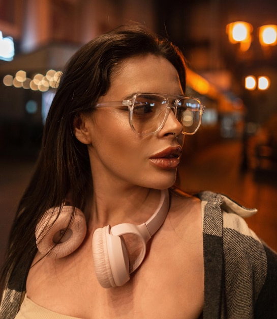 Portrait féminin de rue d'une belle jeune femme hipster avec des lunettes avec un casque rose dans une chemise se promène dans la ville de nuit avec bokeh léger