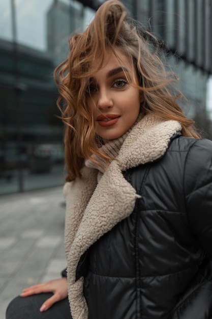 Portrait féminin urbain d'une belle fille heureuse et élégante aux cheveux bouclés dans des vêtements d'hiver à la mode avec un pull tricoté et une veste dans la ville par une journée venteuse