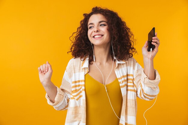 Portrait d'une femme adorable des années 20 portant des écouteurs écoutant de la musique sur un smartphone, isolée sur jaune