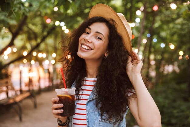 Portrait de femme adorable bouclés portant un chapeau de paille d'été, boire du thé froid dans une tasse en plastique, tout en marchant dans le parc avec des lampes colorées