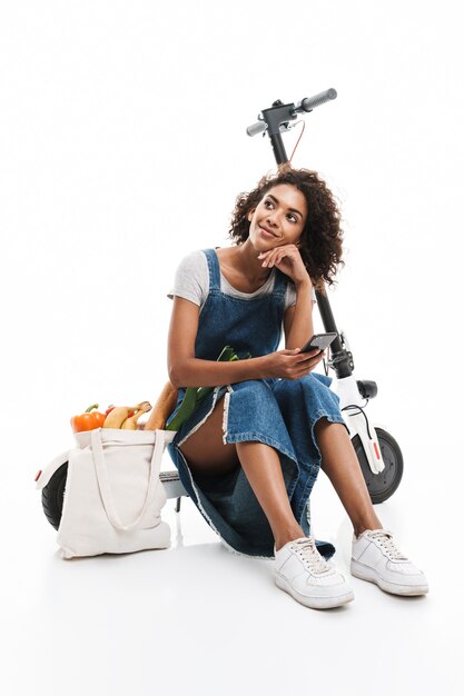 Portrait d'une femme adorable utilisant un téléphone portable alors qu'elle était assise sur un scooter électronique avec un sac d'épicerie isolé sur un mur blanc