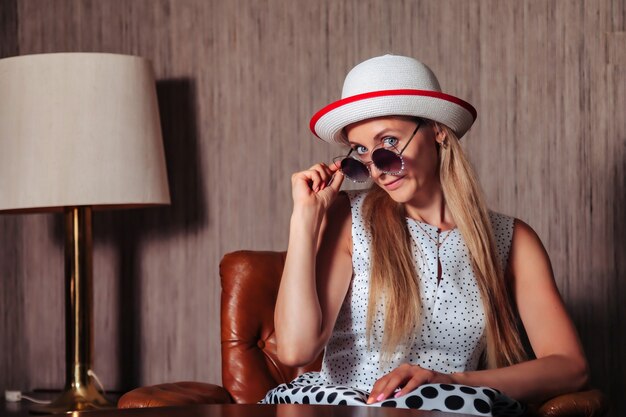 Photo portrait femme adulte actrice posant dans des vêtements de style rétro avec des lunettes de soleil dans le salon vintage. femme en robe noire et blanche de style pin-up à pois et chapeau. concept mode rétro et tendance