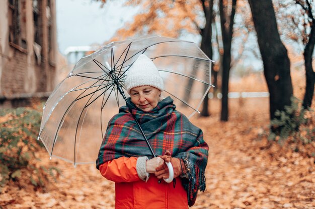 Portrait d'une femme adulte dans un chapeau blanc et une écharpe à carreaux chaude sous un parapluie transparent dans un parc d'automne à l'extérieur. Tapis d'or tombant des feuilles d'automne