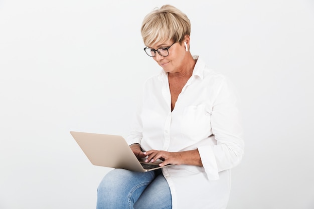 Portrait d'une femme adulte heureuse portant des lunettes et des écouteurs assis avec un ordinateur portable isolé sur un mur blanc en studio