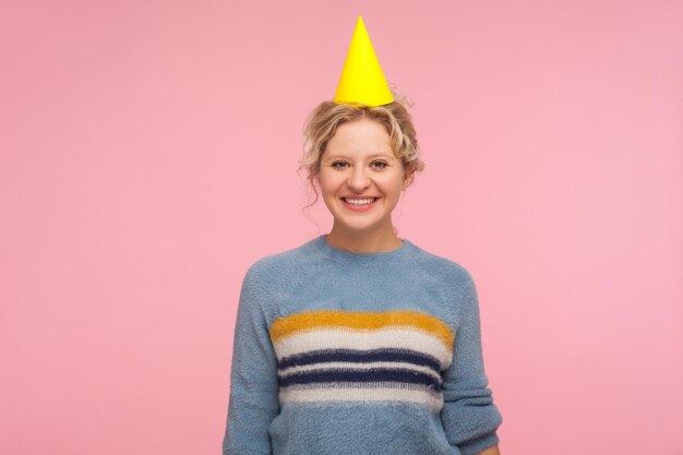 Portrait d'une femme adulte insouciante positive en chandail chaud et chapeau conique drôle sur la tête souriant à la caméra profitant de la fête d'anniversaire célébration des vacances studio intérieur tourné isolé sur fond rose