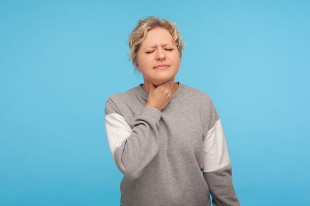 Portrait d'une femme adulte malade et malheureuse aux cheveux bouclés en sweat-shirt touchant le cou souffrant de maux de gorge, de déglutition douloureuse et d'inflammation, de grippe. studio d'intérieur tourné isolé sur fond bleu