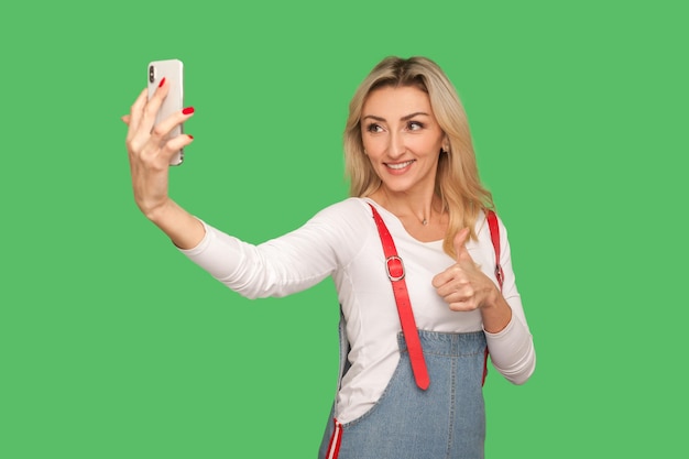 Portrait d'une femme adulte optimiste avec un sourire amical prenant un selfie sur un smartphone et montrant un geste de pouce vers le haut faisant comme un signe d'approbation tout en discutant sur un appel vidéo tourné en studio intérieur isolé