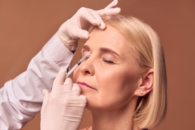 Photo portrait d'une femme adulte plus âgée aux yeux fermés faisant une injection de beauté entre les sourcils les mains dans des gants blancs et une blouse médicale tiennent une seringuelisser les rides