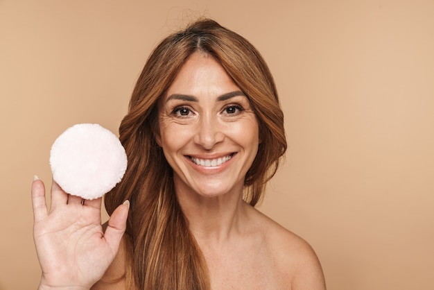 Portrait de femme adulte torse nu à l'aide d'une éponge en poudre et souriant isolé sur beige