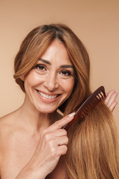 Portrait de femme adulte torse nu se brosser les cheveux et souriant isolé sur beige