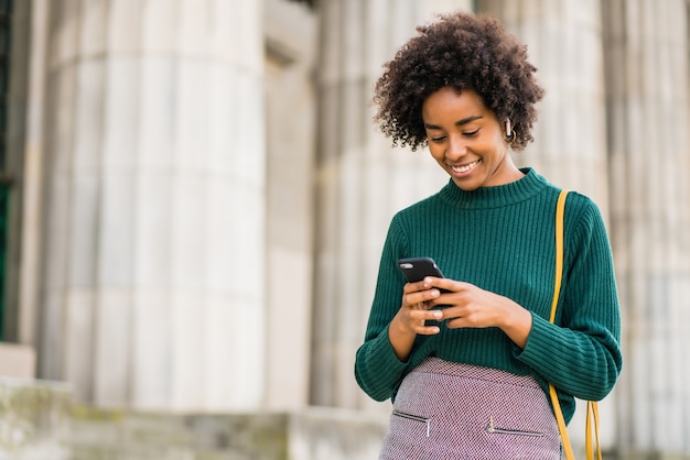 Portrait de femme d'affaires afro à l'aide de son téléphone mobile en se tenant debout à l'extérieur dans la rue