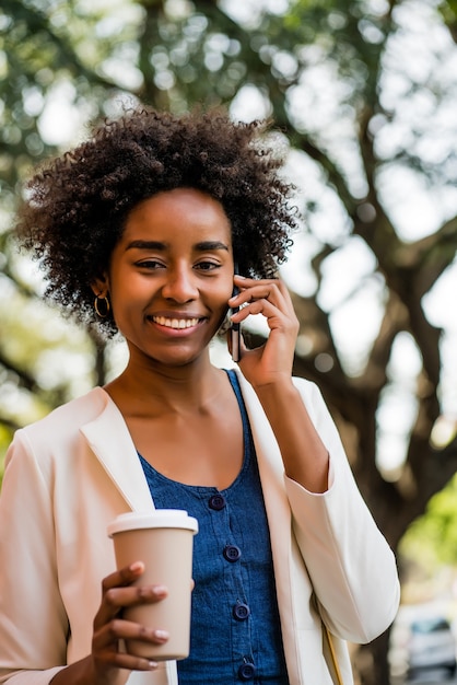 Portrait de femme d'affaires afro parler au téléphone et tenant une tasse de café tout en se tenant à l'extérieur au parc