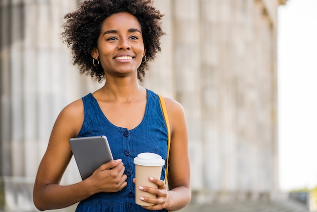 Portrait de femme d'affaires afro tenant une tablette numérique et une tasse de café en se tenant debout à l'extérieur dans la rue