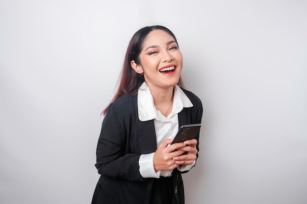 Un portrait d'une femme d'affaires asiatique heureuse sourit et tient son smartphone vêtu d'un costume noir isolé par un fond blanc