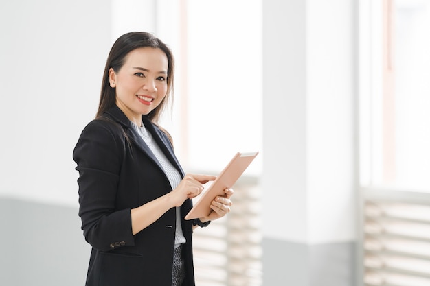 Portrait d'une femme d'affaires asiatique joyeuse et confiante dans un costume d'affaires debout à l'aide d'une tablette numérique dans le bâtiment de l'entreprise. Banque d'images d'affaires