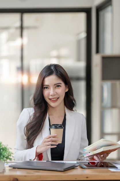 Portrait d'une femme d'affaires asiatique séduisante tenant une tasse de café et un cahier assis au bureau souriant et regardant la caméra