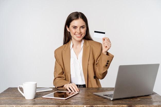 Portrait d'une femme d'affaires assise au bureau, montrant une carte de crédit et souriante, travaillant à table avec un ordinateur portable et une tablette, fond blanc.