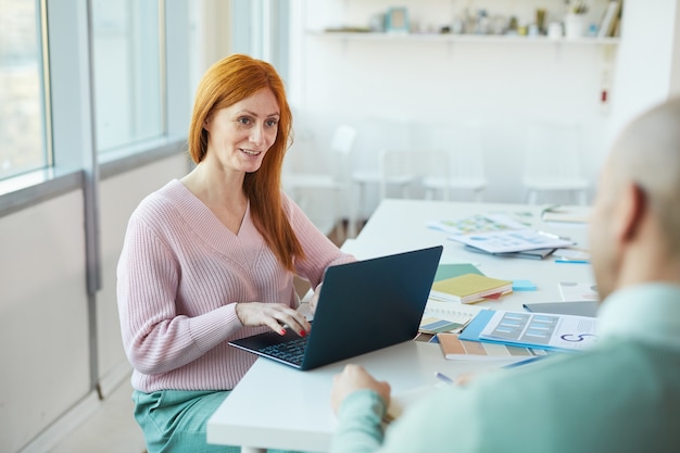 Portrait de femme d'affaires aux cheveux rouges matures interviewant l'homme tout en utilisant un ordinateur portable au bureau, copiez l'espace