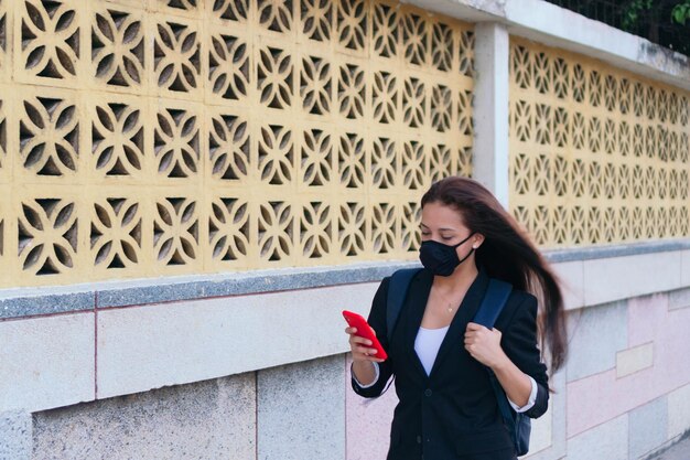 Portrait d'une femme d'affaires belle et prospère portant un masque de protection et tenant un smartphone en marchant dans la rue.