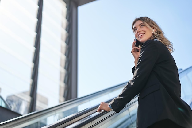 Portrait de femme d'affaires en costume noir qui monte sur l'escalator en parlant sur la vendeuse de téléphone mobile wal