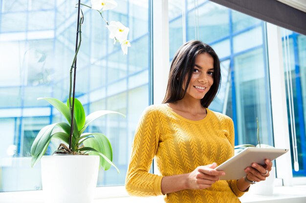 Portrait d'une femme d'affaires décontractée souriante debout avec une tablette au bureau près de la fenêtre