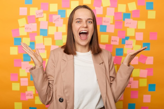 Portrait d'une femme d'affaires extrêmement heureuse excitée portant une veste beige debout contre un mur jaune avec des cartes mémo colorées criant de bonheur les bras levés a des affaires prospères