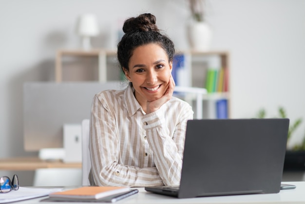 Portrait d'une femme d'affaires heureuse assise sur son lieu de travail devant un ordinateur portable et souriant à la caméra à